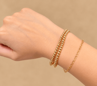 A close-up of a person's wrist wearing three gold bracelets: two beaded bangle bracelets and one delicate chain bracelet, against a soft beige background.
