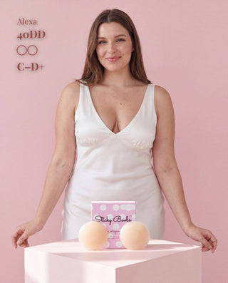 A woman in a white satin slip dress stands behind a pink pedestal displaying two beige sticky boobs and a pink box labeled 'Sticky Boobs'. Text on the top left reads 'Alexa 40DD C-D+'.