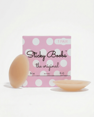 A pink polka-dot box labeled 'Sticky Boobs the original' with two flesh-toned adhesive nipple covers placed in front on a clean white background.