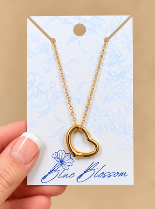 A gold open heart pendant necklace on a delicate gold chain, displayed on a white card with blue floral patterns and the brand name 'Blue Blossom'. A hand with a French manicure is holding the card on the left side, set against a solid beige background.