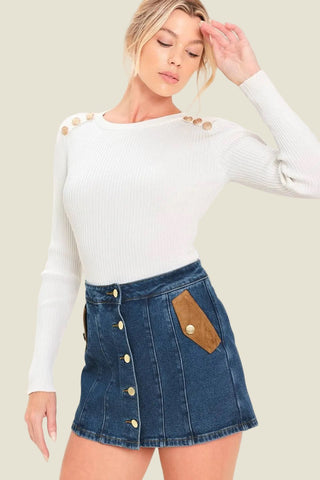 A woman modeling a white ribbed long-sleeve top with gold button details on the shoulders and a blue denim mini skirt with gold buttons and a brown suede pocket, against a plain light background.