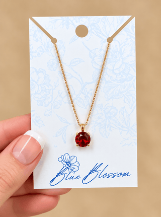 A gold chain necklace with a round red gemstone pendant, displayed on a white card with blue floral details and the brand name 'Blue Blossom'. A hand with a French manicure is holding the card against a solid beige background.