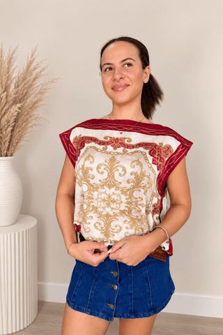 A woman wearing a sleeveless top with a cream and gold damask pattern and red trim, paired with denim shorts, standing in a brightly lit room with a neutral wall and a potted dried grass arrangement.