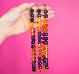 Three beaded fringe necklaces with alternating translucent orange and dark purple beads, held up by a hand against a solid pink background.