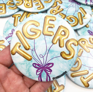 Button badge with 'TIGERS' in gold letters and a purple bow design on a light blue background.