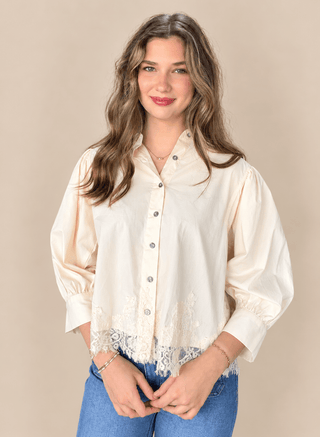 A woman modeling a cream-colored long-sleeve blouse with lace trim at the hem and button-front closure, paired with blue jeans against a plain beige studio background.