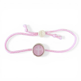 A light pink braided cord bracelet featuring a round pink medallion with a white cross, set in a silver-toned bezel, and a small white wooden bead with a cross on the adjustable sliding knot.