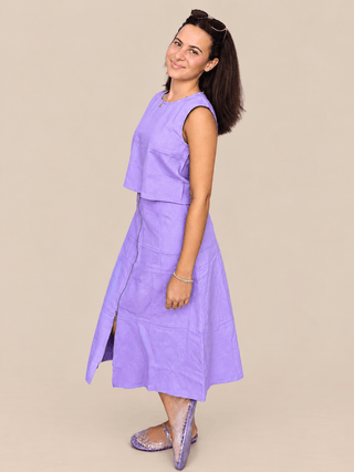 A woman modeling a lavender sleeveless midi dress with a tiered ruffle top and side slit, paired with patterned flats, against a solid beige background.