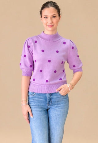 A woman modeling a lavender purple mock neck knit sweater with dark purple polka dots and puff sleeves, paired with blue jeans, against a solid beige background.