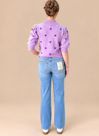 A woman shown from the back wearing a lavender purple knit sweater with dark purple polka dots and light blue jeans, standing against a solid beige studio background.