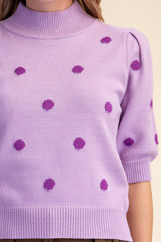 A close-up of a lavender knit sweater with darker purple textured polka dots, featuring a mock neck and short puff sleeves, shown on a model against a light beige background.