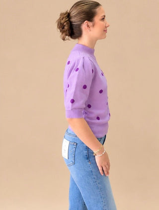 A woman in side profile wearing a lavender purple short-sleeve knit sweater with darker purple polka dots and light blue jeans, set against a solid beige studio background.