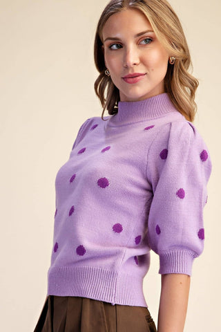 A woman wearing a light purple knit mock-neck sweater with darker purple pom-pom dots, paired with a brown skirt, against a solid beige background.
