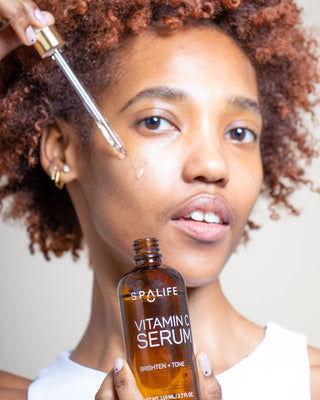 Woman applying vitamin C serum to her face with a dropper, holding the bottle labeled 'SpaLife'.