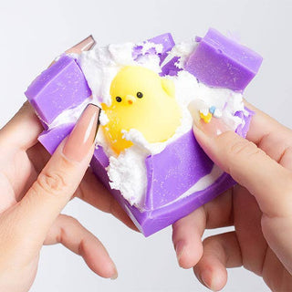 A person's hands holding a purple and white textured bar of soap that is cracked open to reveal a yellow rubber chick toy inside, set against a plain light gray background.