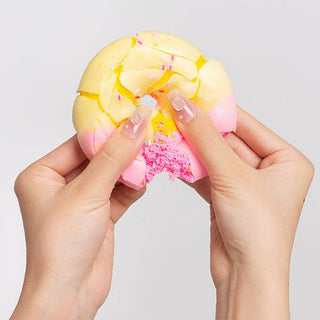 A close-up of two hands holding and crushing a pastel yellow and pink squishy toy with a cracked, broken surface and small pink sprinkles, against a light gray background.