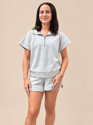 A woman modeling a light gray heathered short-sleeve half-zip pullover and matching shorts against a solid beige studio background.