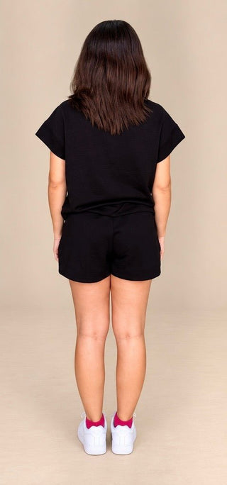 A child model shown from the back wearing a black short-sleeve t-shirt and matching black shorts, paired with white sneakers and pink socks, against a solid beige studio background.