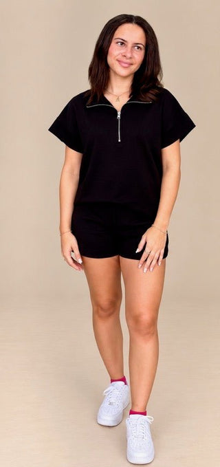 A woman modeling a black short-sleeve half-zip pullover top with matching black shorts, styled with white sneakers, against a solid beige studio background.