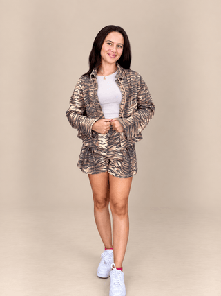 A woman modeling a matching metallic tiger-print jacket and shorts set with a white top and white sneakers, standing against a solid beige studio background.